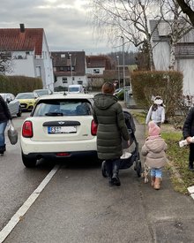 Zu-Fuß-Gehende quetschen sich auf dem restlichen Gehweg, im Vorgarten eines Hauses und auf der Fahrbahn an einem auf dem Gehweg geparkten Auto vorbei.